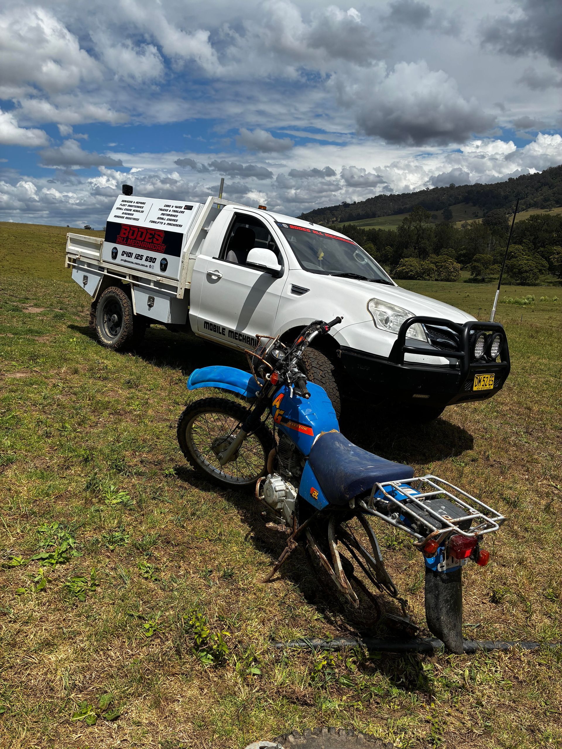 White truck and blue motorcycle parked on a grassy hill, cloudy sky in the background. — Bodes All Mechanical In Bega, NSW