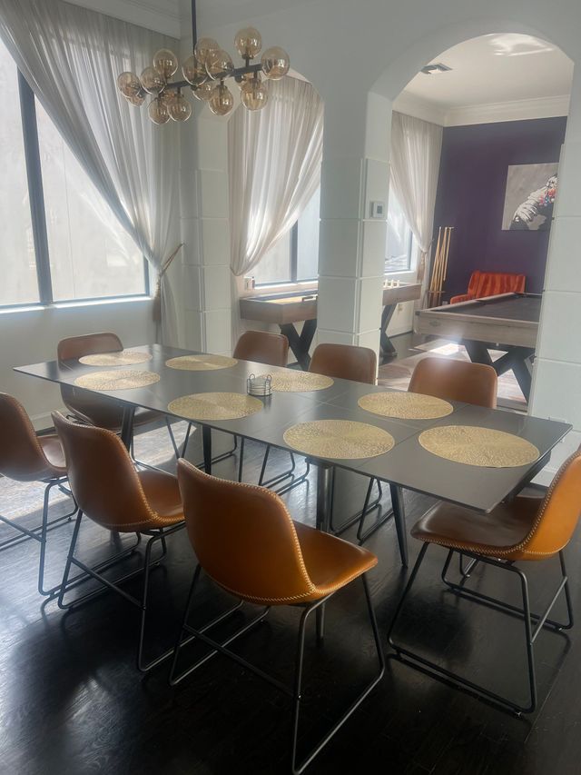 Dining room with long table, tan chairs, sheer curtains, and chandelier in soft natural light