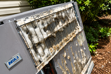 Broken white window air conditioner with rust and grime, lying outdoors near bushes.