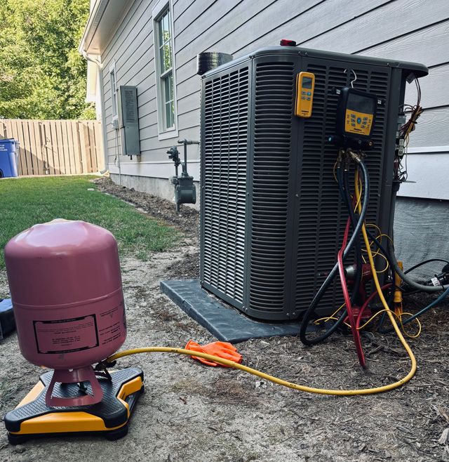 Outdoor HVAC unit with gauges and hoses attached next to a house, and a pink tank on a dolly in the foreground
