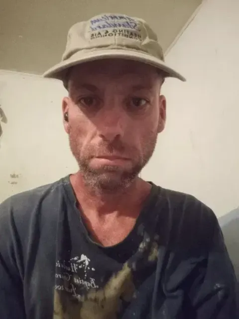 Man in a hat and dark shirt, looking directly at the camera with a serious expression.