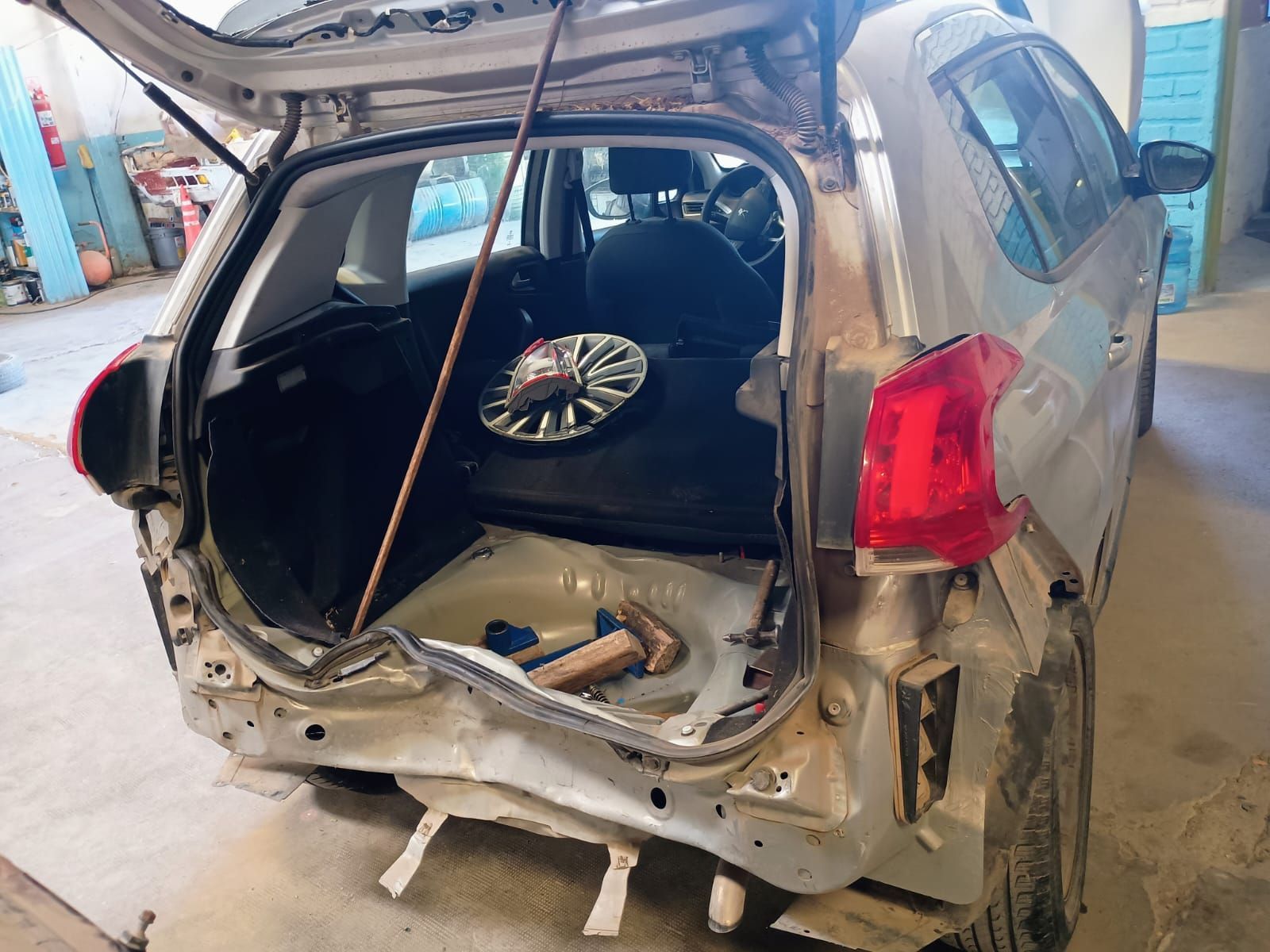 Automóvil plateado dañado con el maletero abierto en un taller de reparación; muestra daños en la carrocería trasera.