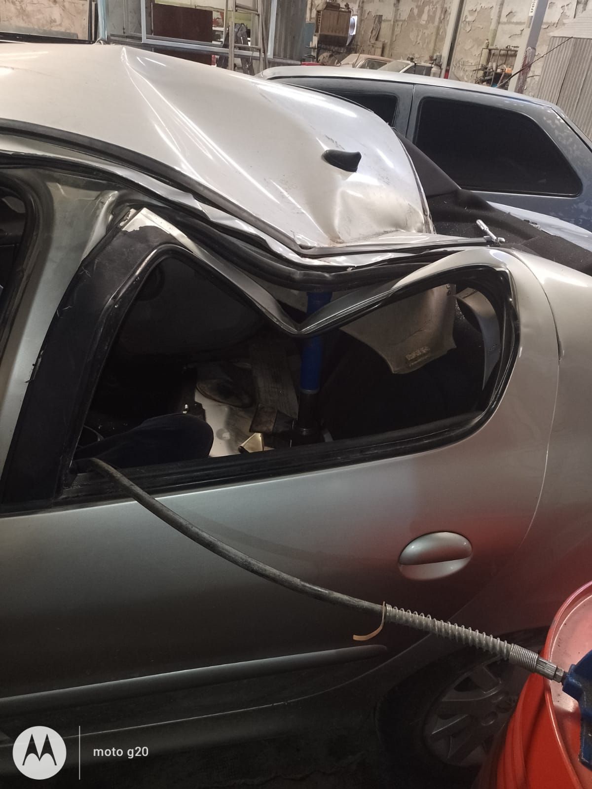 Auto plateado dañado, con el techo destrozado y la ventana rota, posiblemente en un taller.