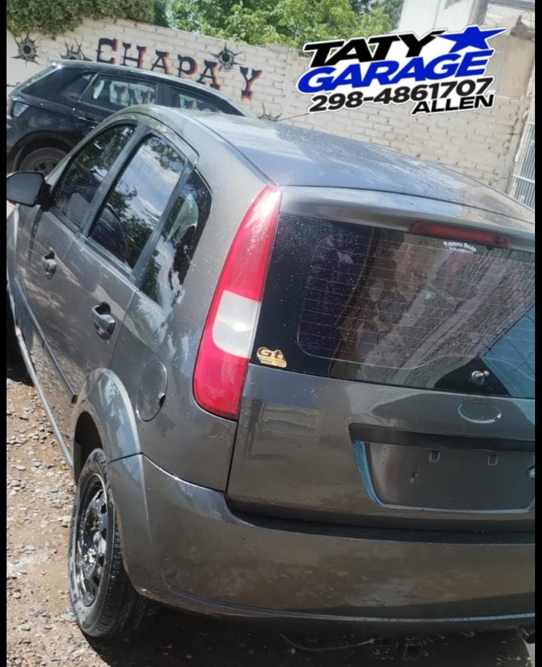 Ford Fiesta hatchback gris estacionado cerca de un garaje con señalización comercial.