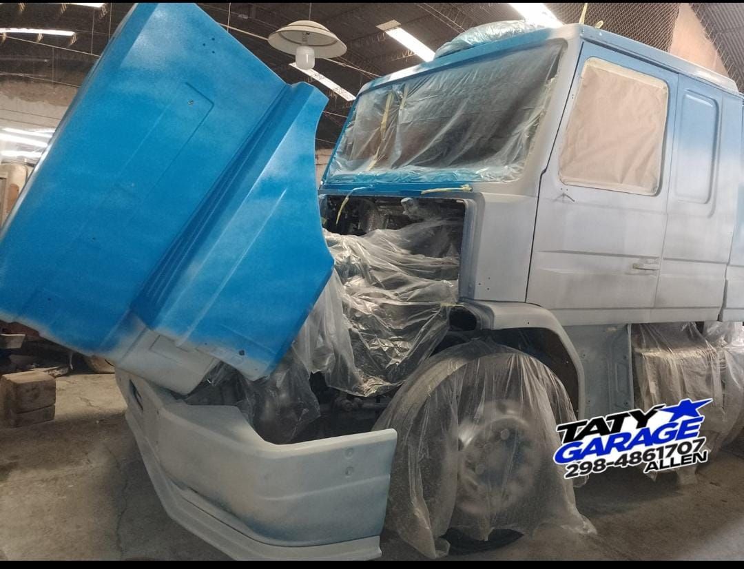 Cabina de camión pintada de azul en un garaje, con el capó levantado y partes cubiertas de plástico.