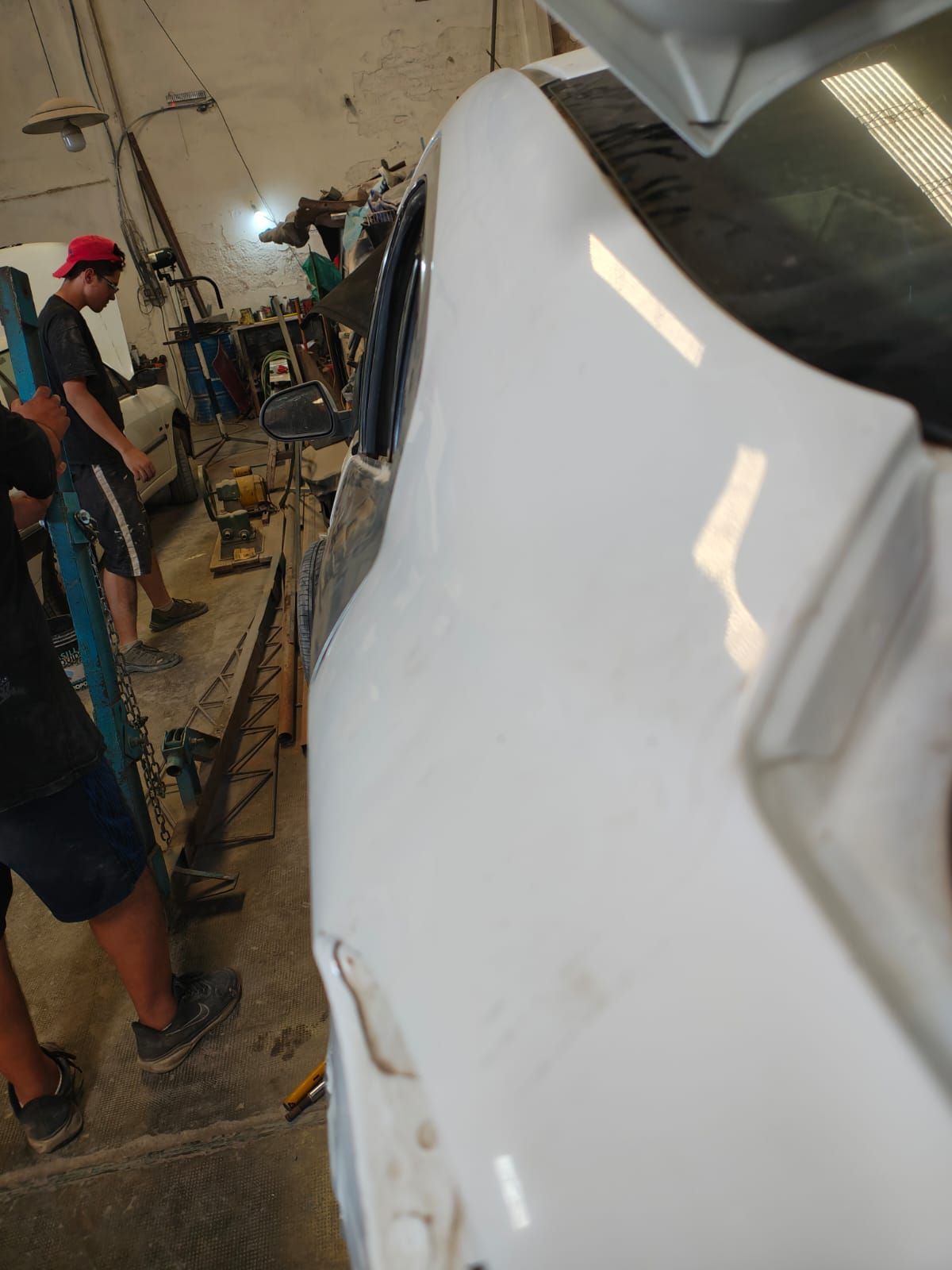 Se está reparando un coche blanco en un taller; se ven dos personas, una de ellas con un sombrero rojo.