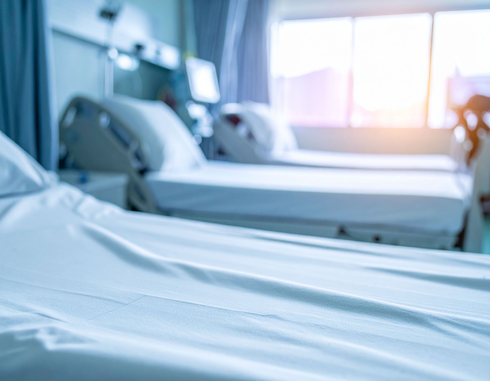 Hospital beds in a bright room, suggesting a recovery setting. The beds are empty with clean white sheets.