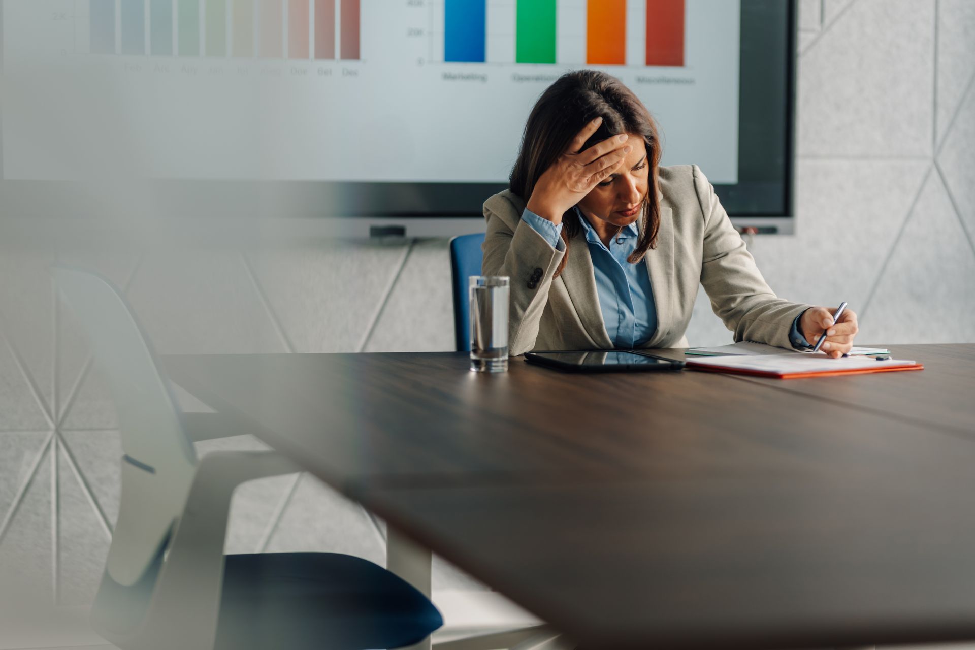 Woman sitting at a conference table, holding her head and writing notes in an office meeting room.
