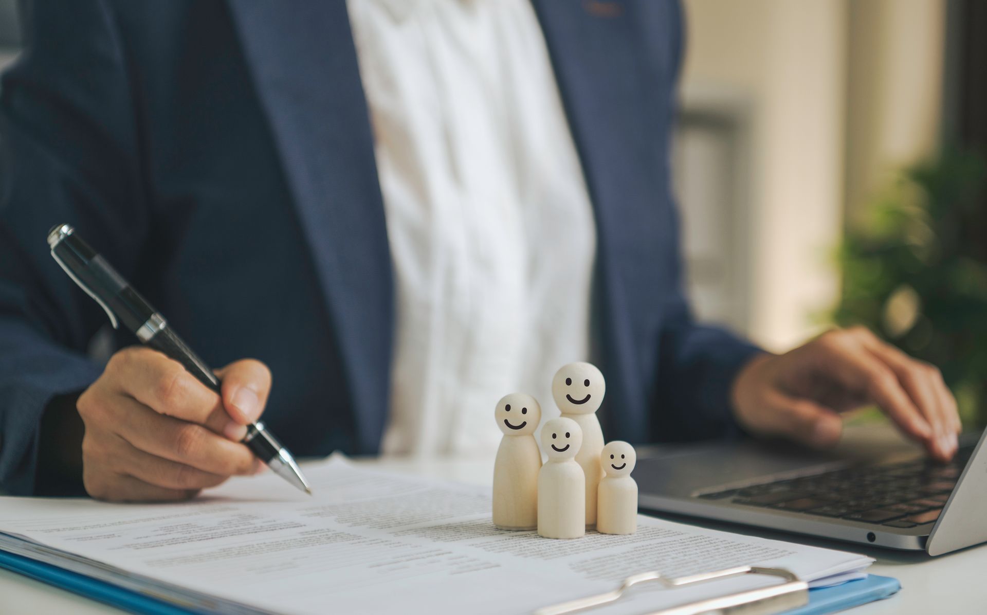 Businessperson signing a document beside a laptop, with small smiling wooden figures on the desk