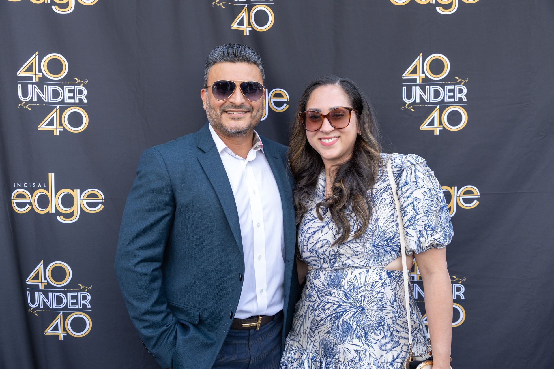 Two people posing at a “40 Under 40” event backdrop, smiling in sunglasses and business attire