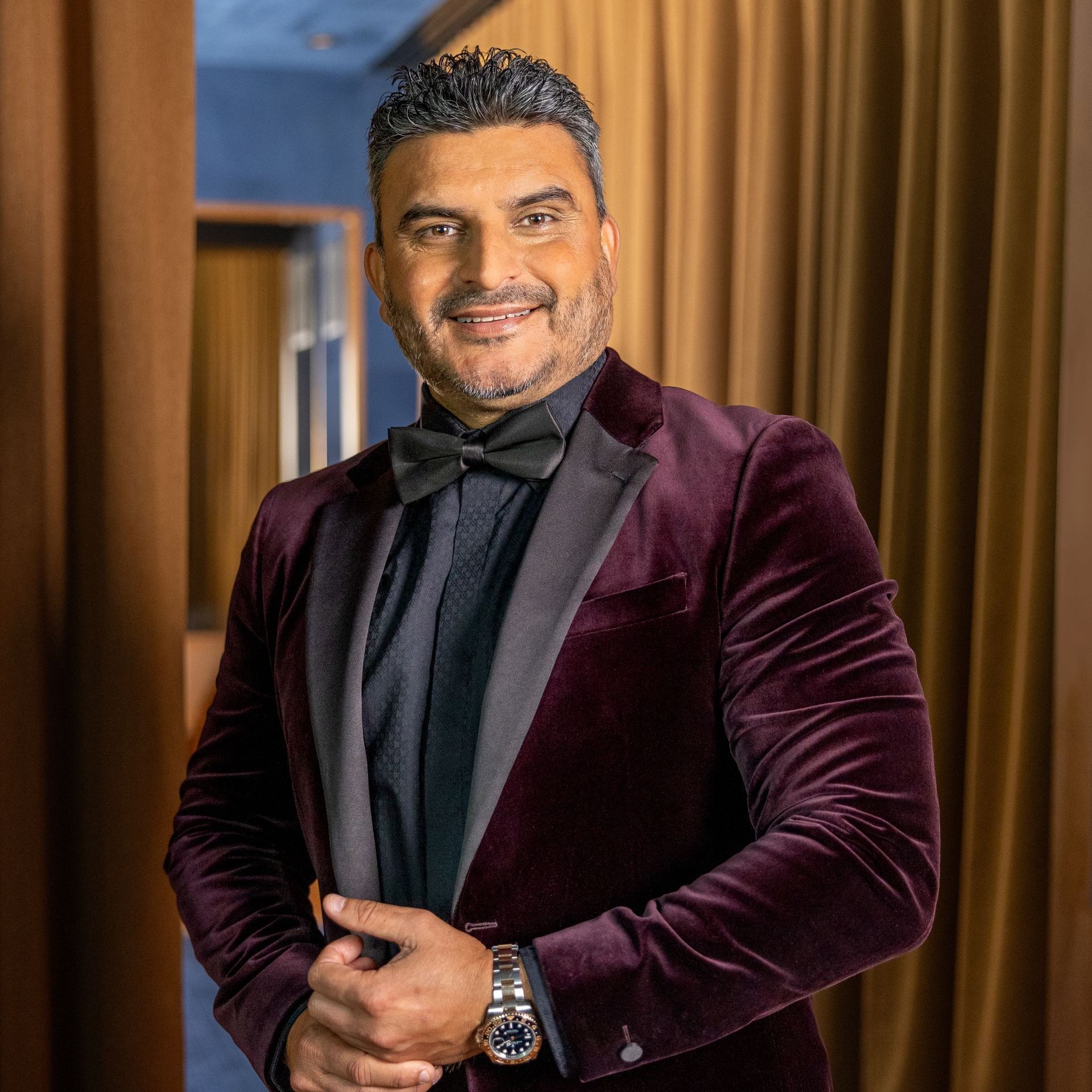 Man in a burgundy velvet blazer and black bow tie, smiling in a warm indoor setting.