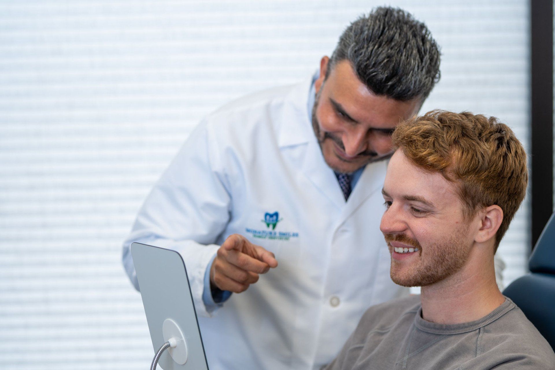 Dentist smiling and pointing at a patient’s teeth in a bright clinic, with a mirror in the foreground