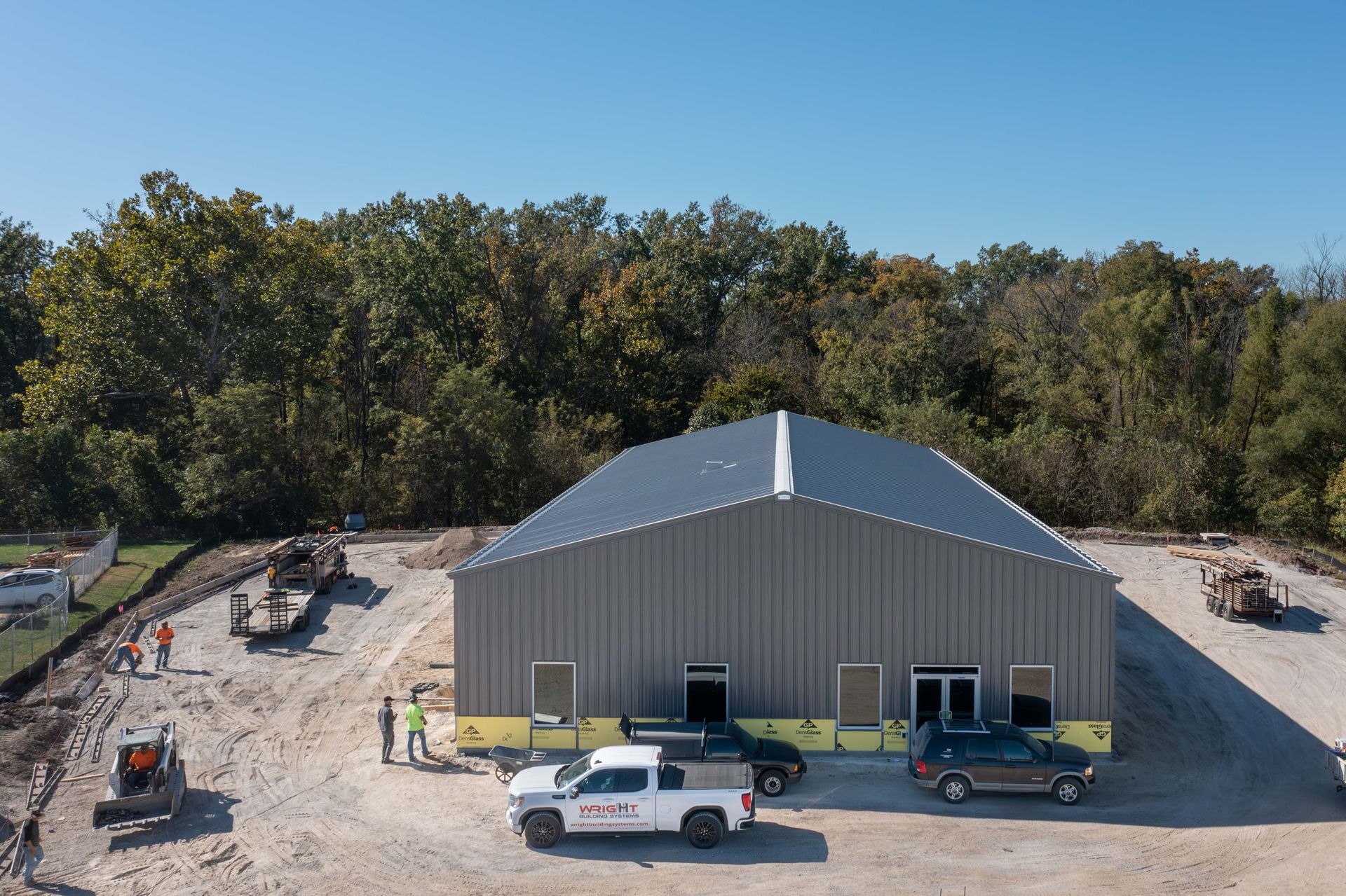 Large gray metal building under construction with work trucks and equipment in front, trees in the background