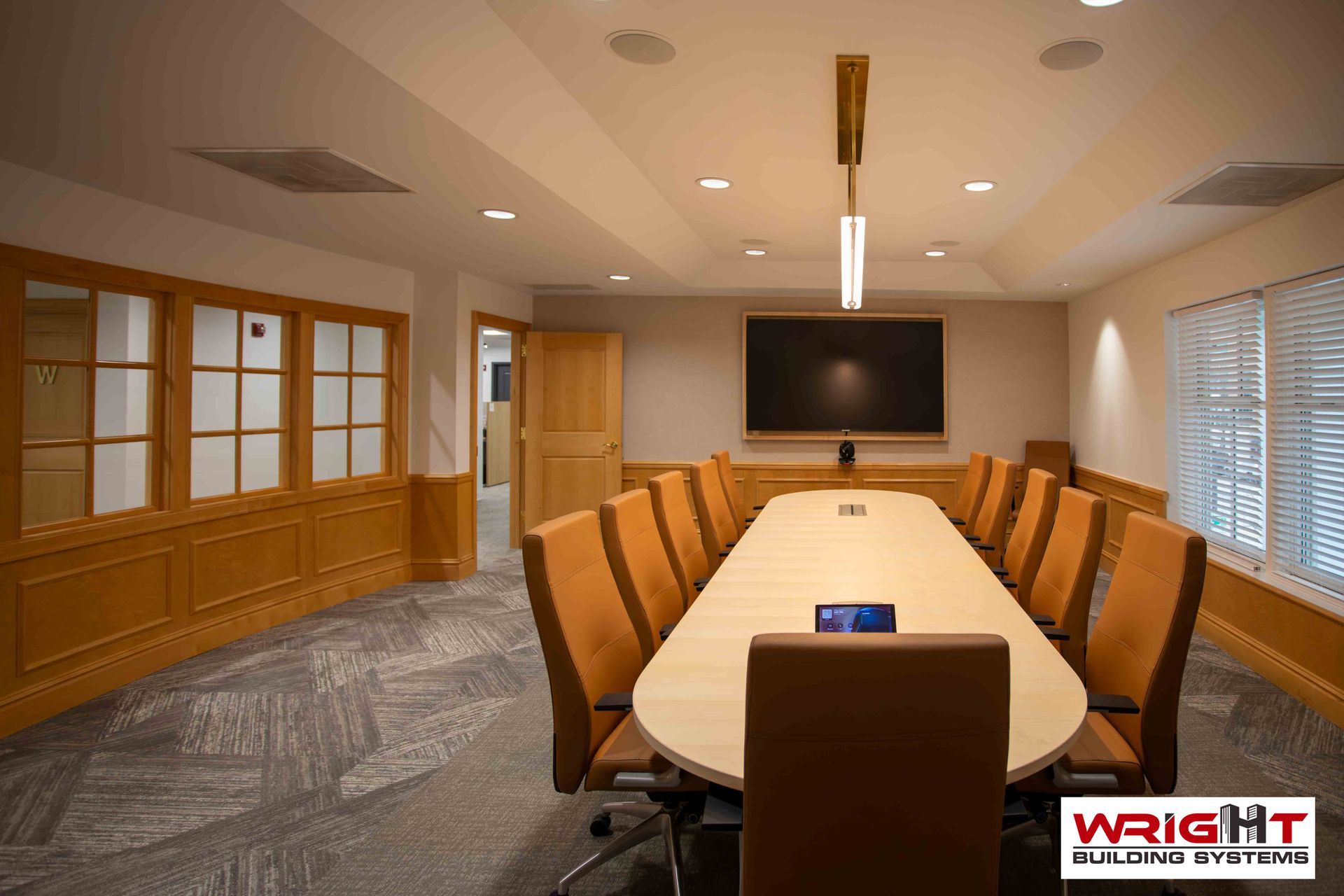 A conference room with a long oval table surrounded by orange chairs, wooden paneling, a wall-mounted screen, and blinds.