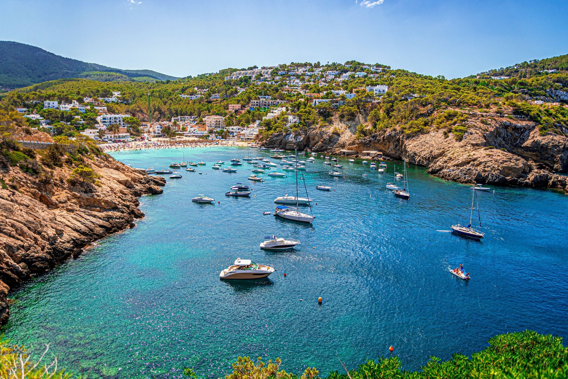 Bay with boats, clear blue water, beach, and buildings on a hillside under a sunny sky.