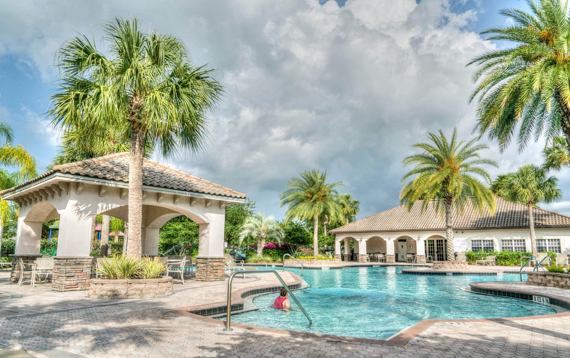 Swimming pool with palm trees, gazebo, and cloudy sky. A person swims in the water.