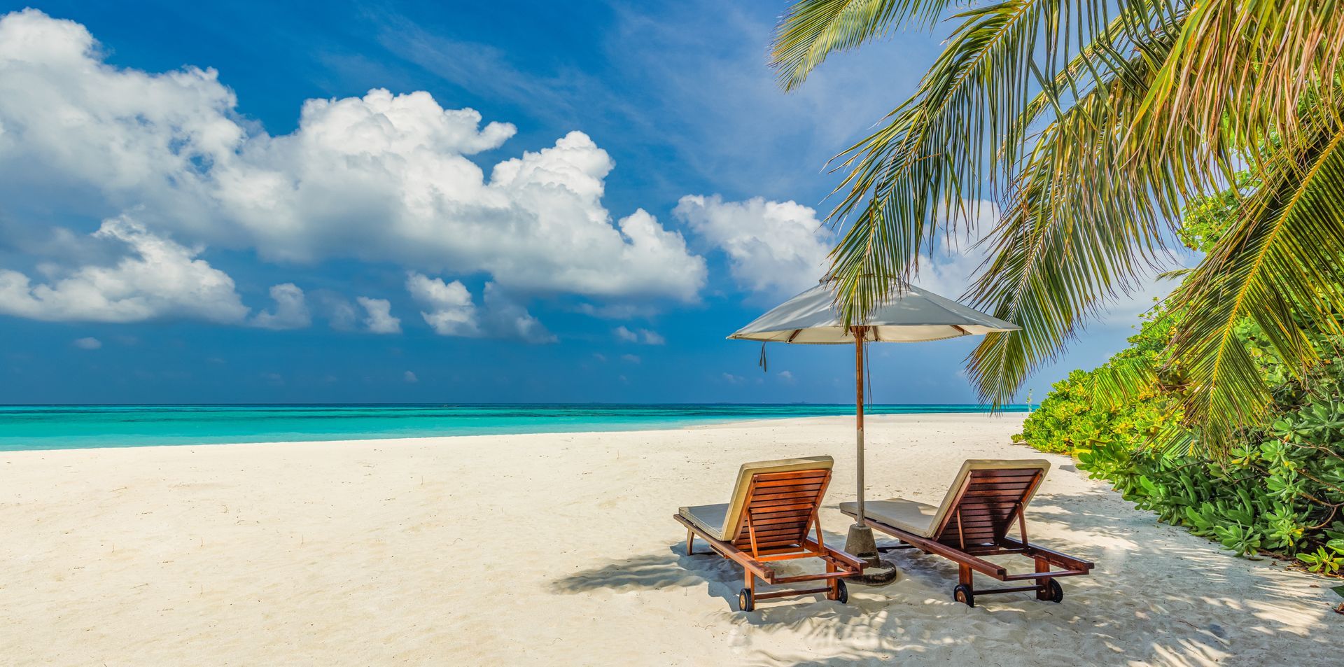 Tropical beach scene with white sand, turquoise water, palm trees, two lounge chairs, and an umbrella.