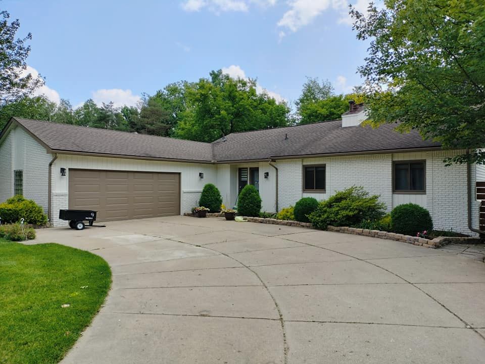 White brick house with brown roof, attached garage, and circular driveway. Green bushes and trees surround the property.
