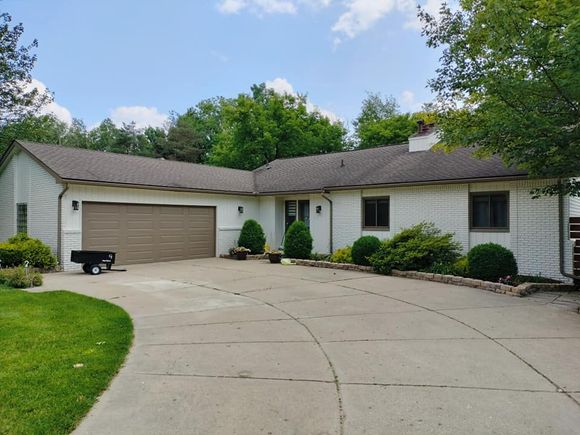 White brick house with brown roof, attached garage, and circular driveway. Green bushes and trees surround the property.