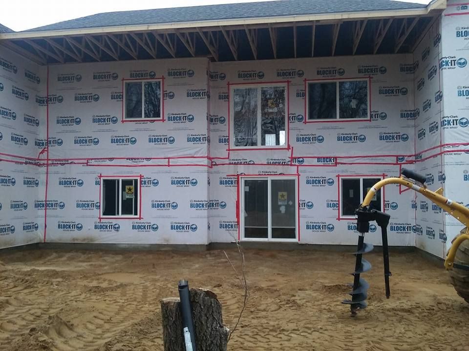 Exterior of a house under construction; windows and door framed, wrapped in blue protective material. Auger in foreground.