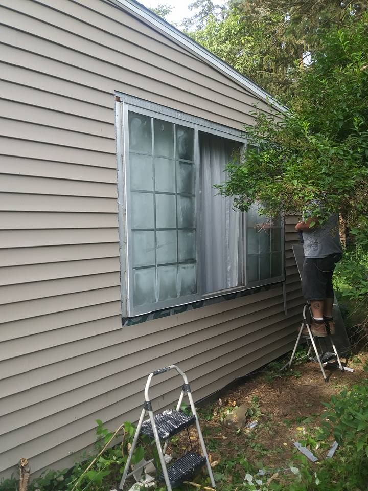 Person on ladder working on a window of a house with light brown siding; another small ladder is at the bottom.