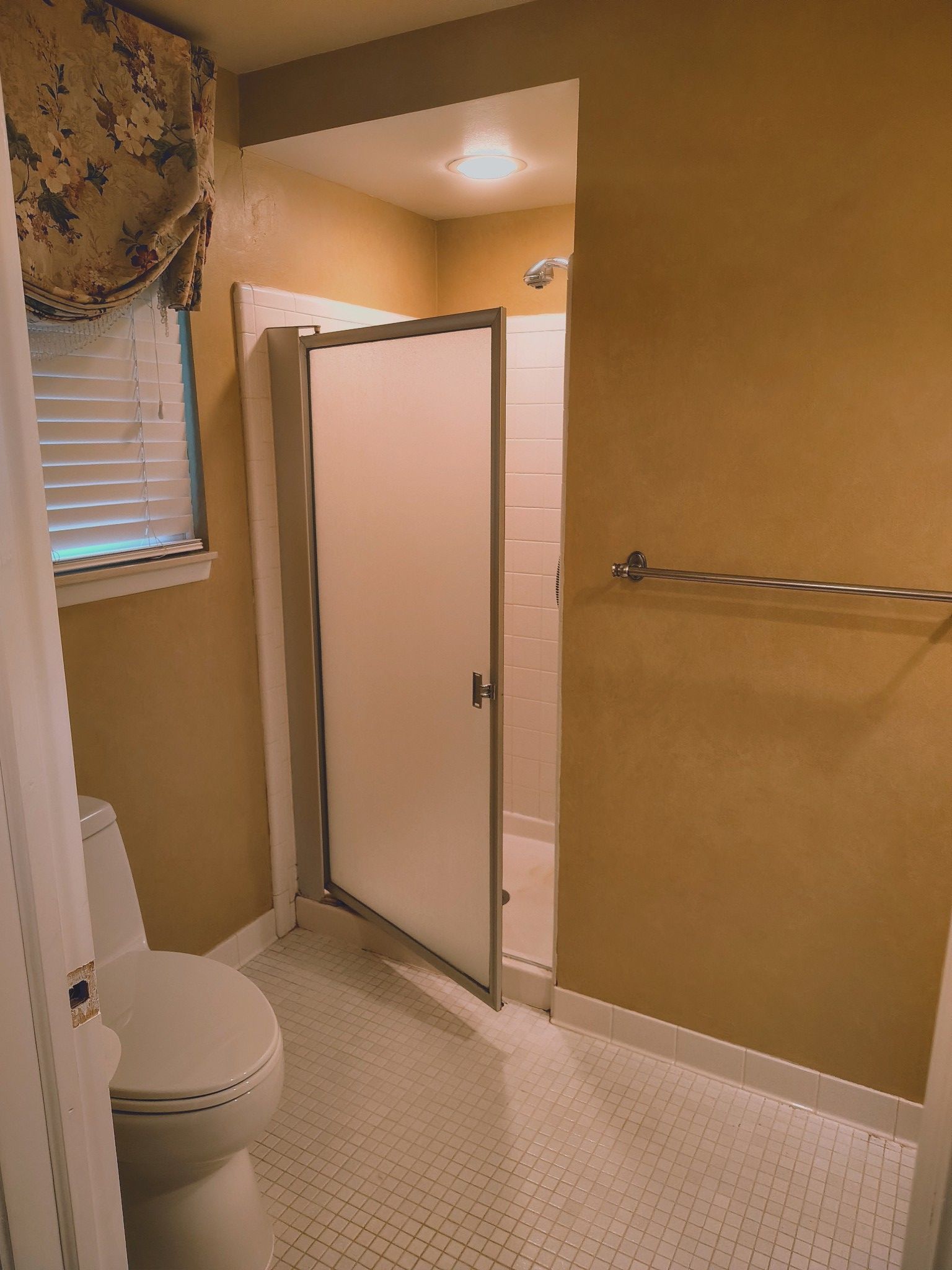 Bathroom interior with toilet, shower, and window with blinds. Yellow walls, white tile floor.