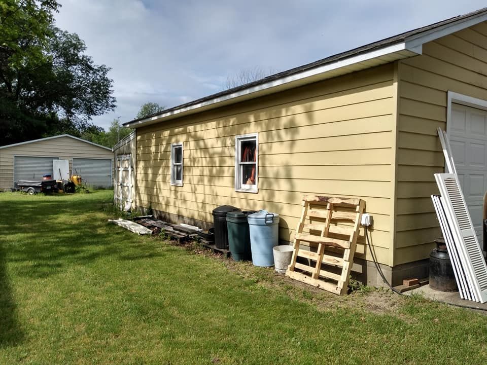 Yellow-sided garage next to a green lawn with a person looking out a window. Trash cans and pallets are visible.