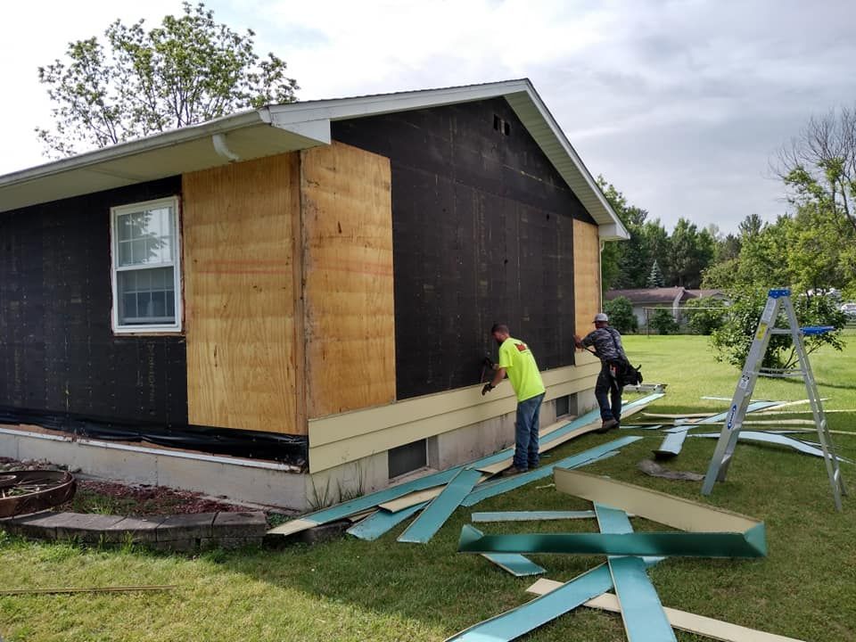 Two workers install siding on a house exterior; tan plywood visible, green siding strips on grass.