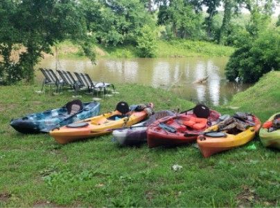 Colorful kayaks lined up on grass beside a calm river, with chairs under trees in the background