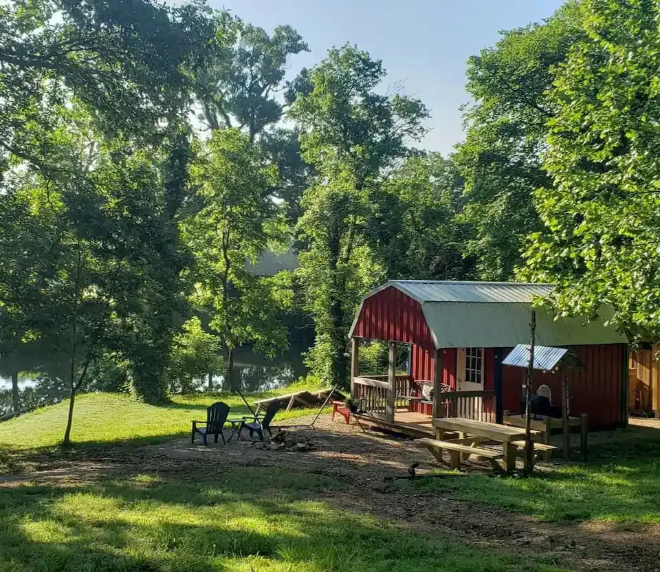 Red cabin in a sunny wooded clearing with picnic tables and chairs near a lake