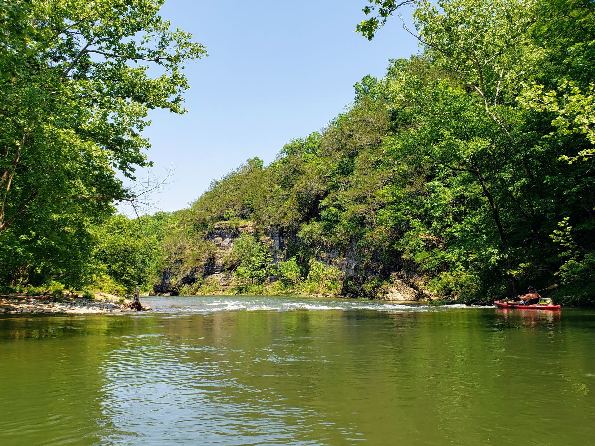Calm green river bordered by dense trees and rocky cliffs under a clear blue sky