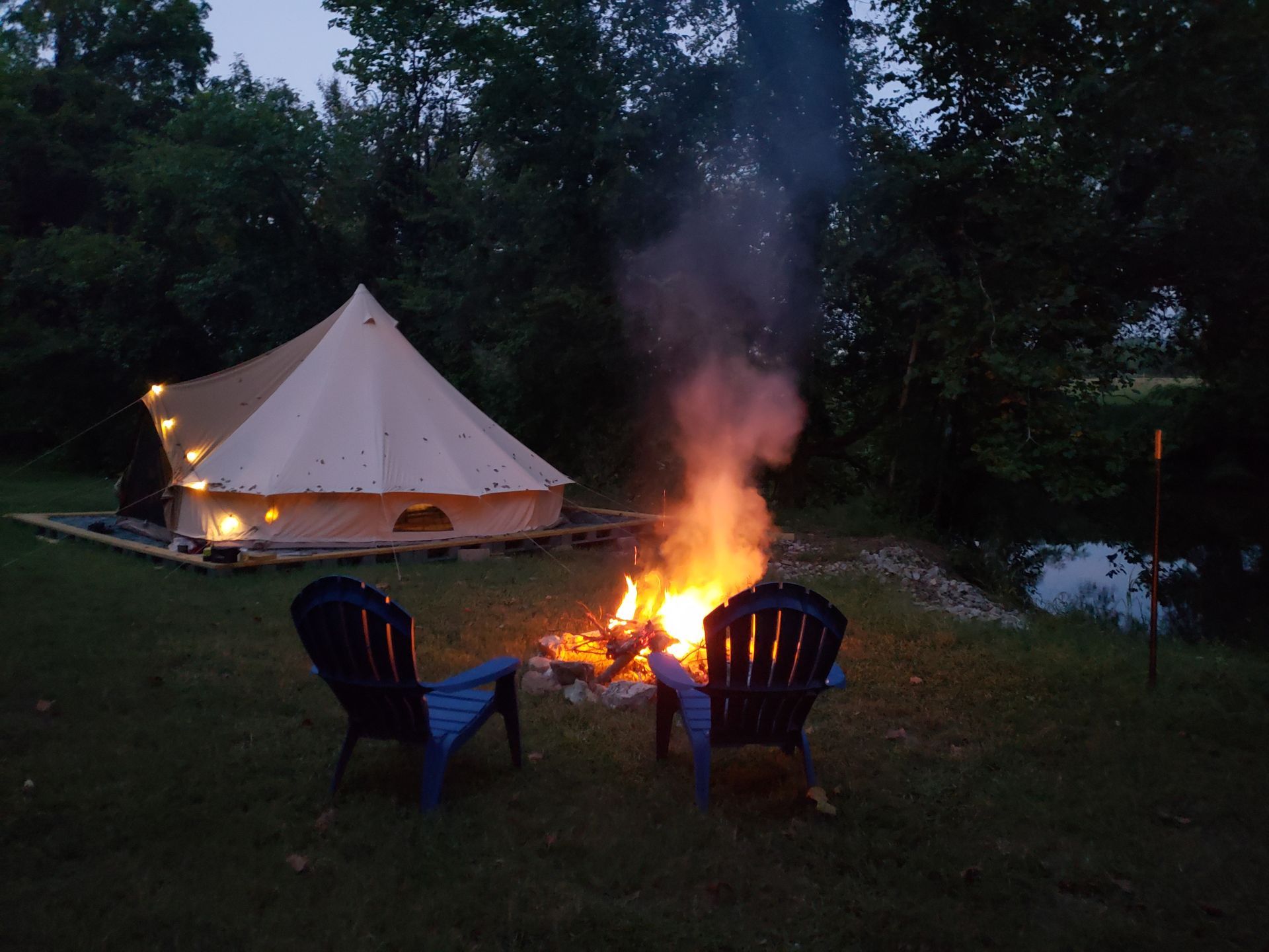 Canvas tent and campfire at dusk with two blue chairs in a wooded campsite