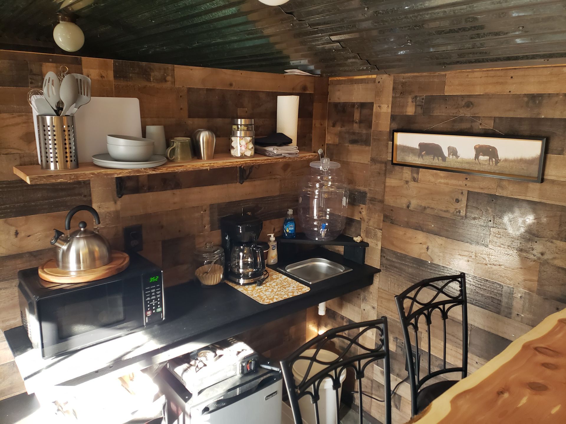 Rustic kitchen with wood walls, black counter, metal kettle, dishes, and dining chairs.