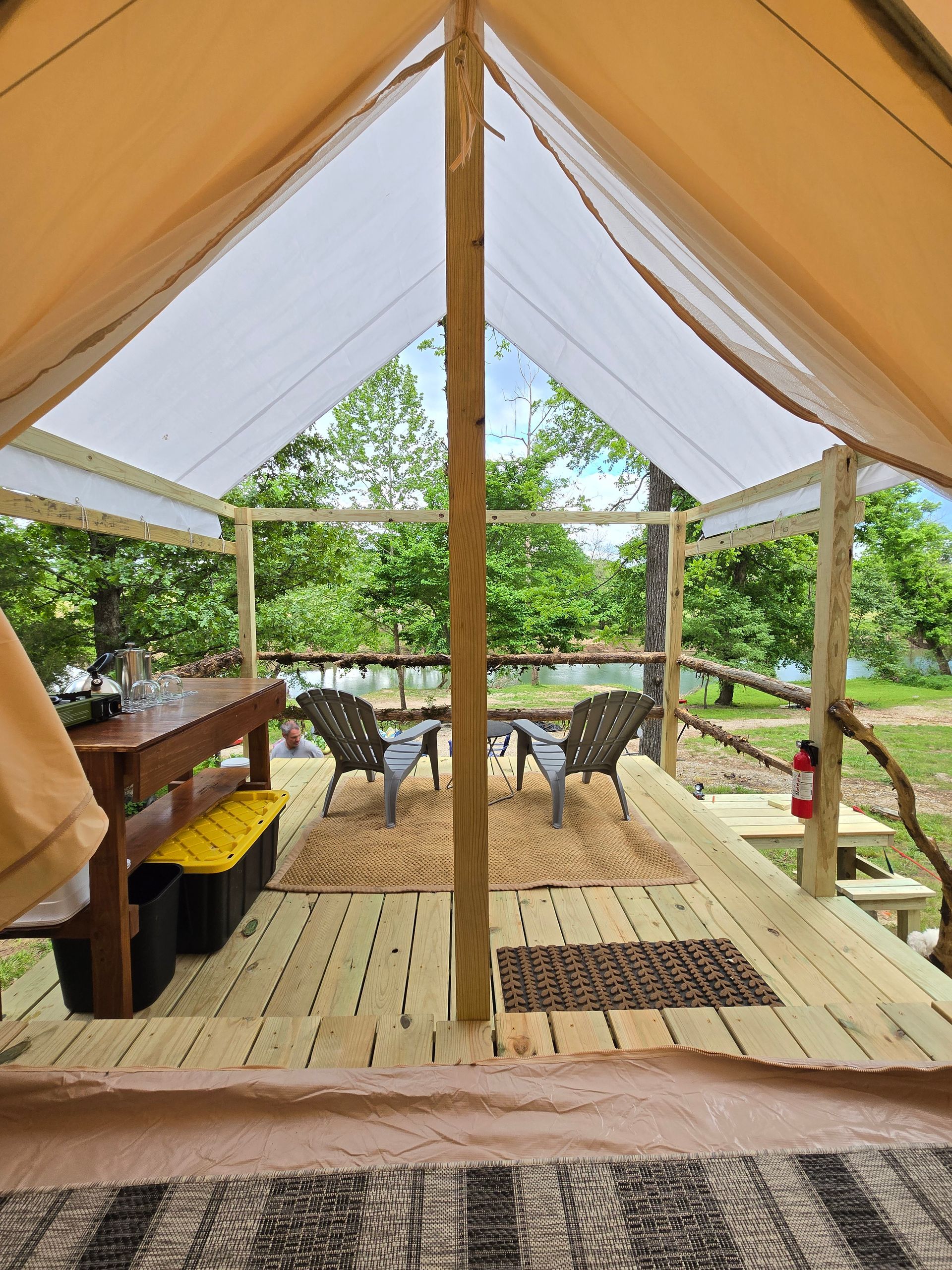 Open-air lounge tent with wooden deck, chairs, and table overlooking green trees
