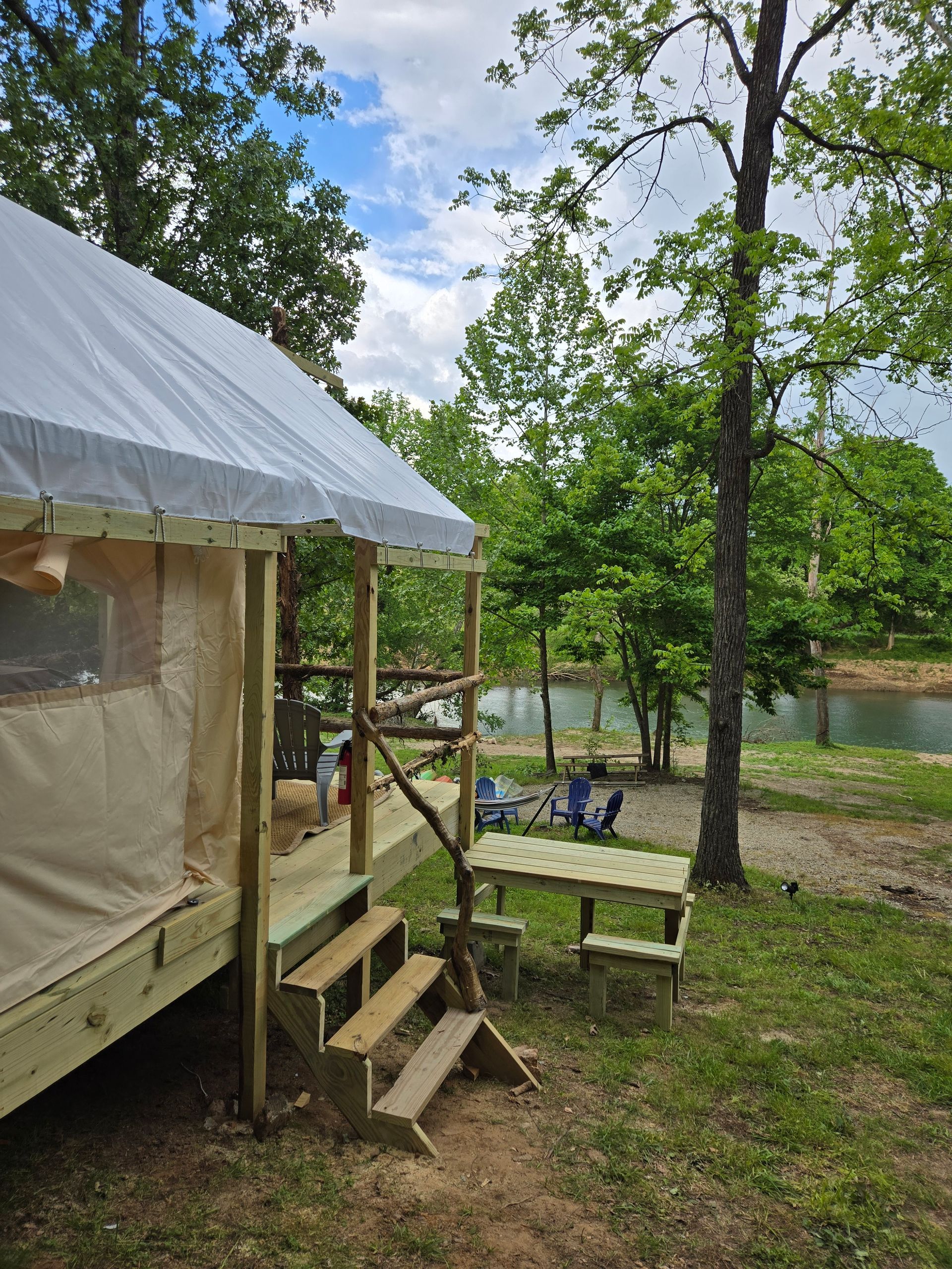 Canvas tent with wooden steps and chairs beside a wooded lakeside campsite