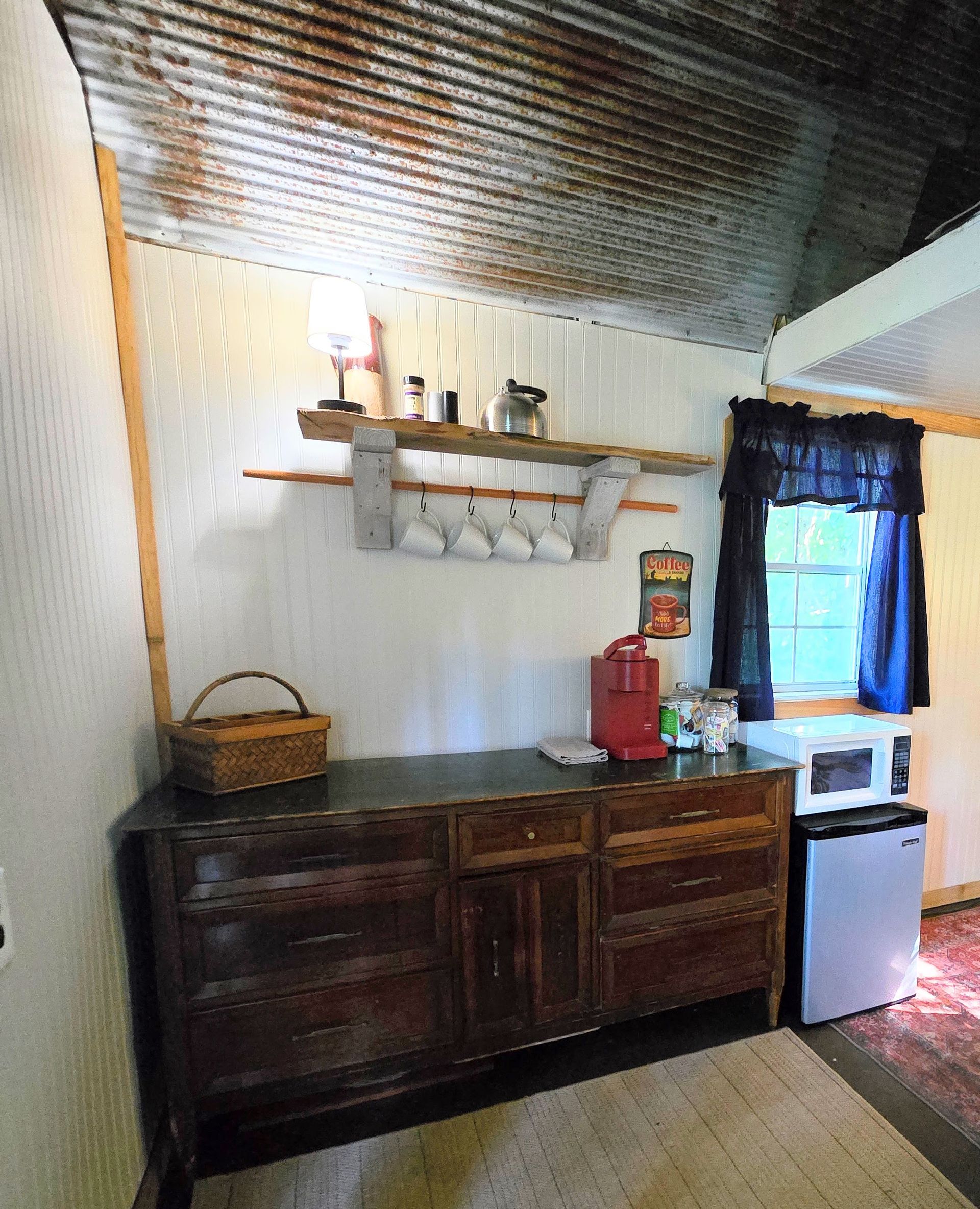 Rustic kitchen with dark wood cabinets, shelves, basket, and a small white refrigerator by a window.