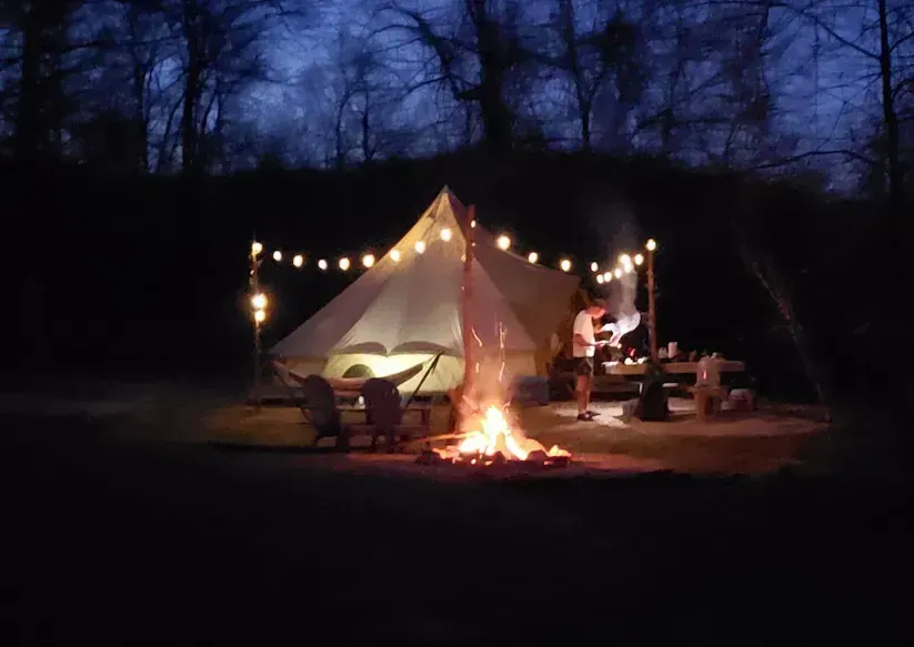Lit camping tent with string lights and a campfire at dusk in a wooded clearing
