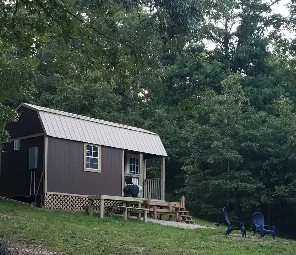 Small dark cabin with porch in a wooded yard, with two blue chairs on the grass.