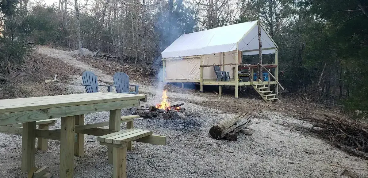 Wooden picnic table and bench by a campfire near a small cabin in a wooded outdoor area