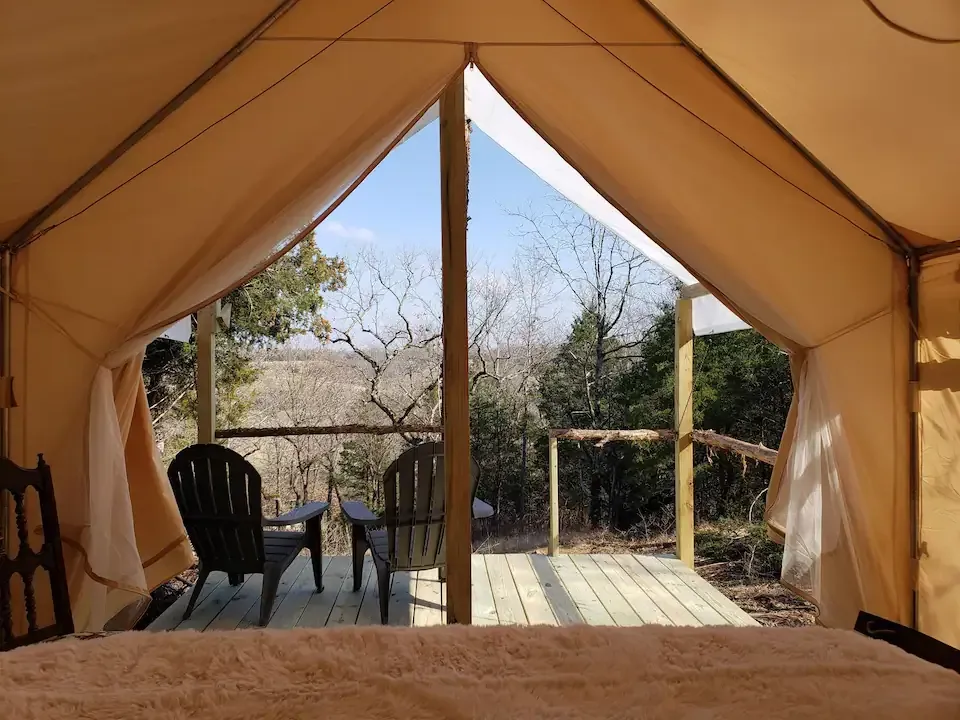 Inside a tan tent, a patio chair and small table overlook a wooden deck and leafless trees.
