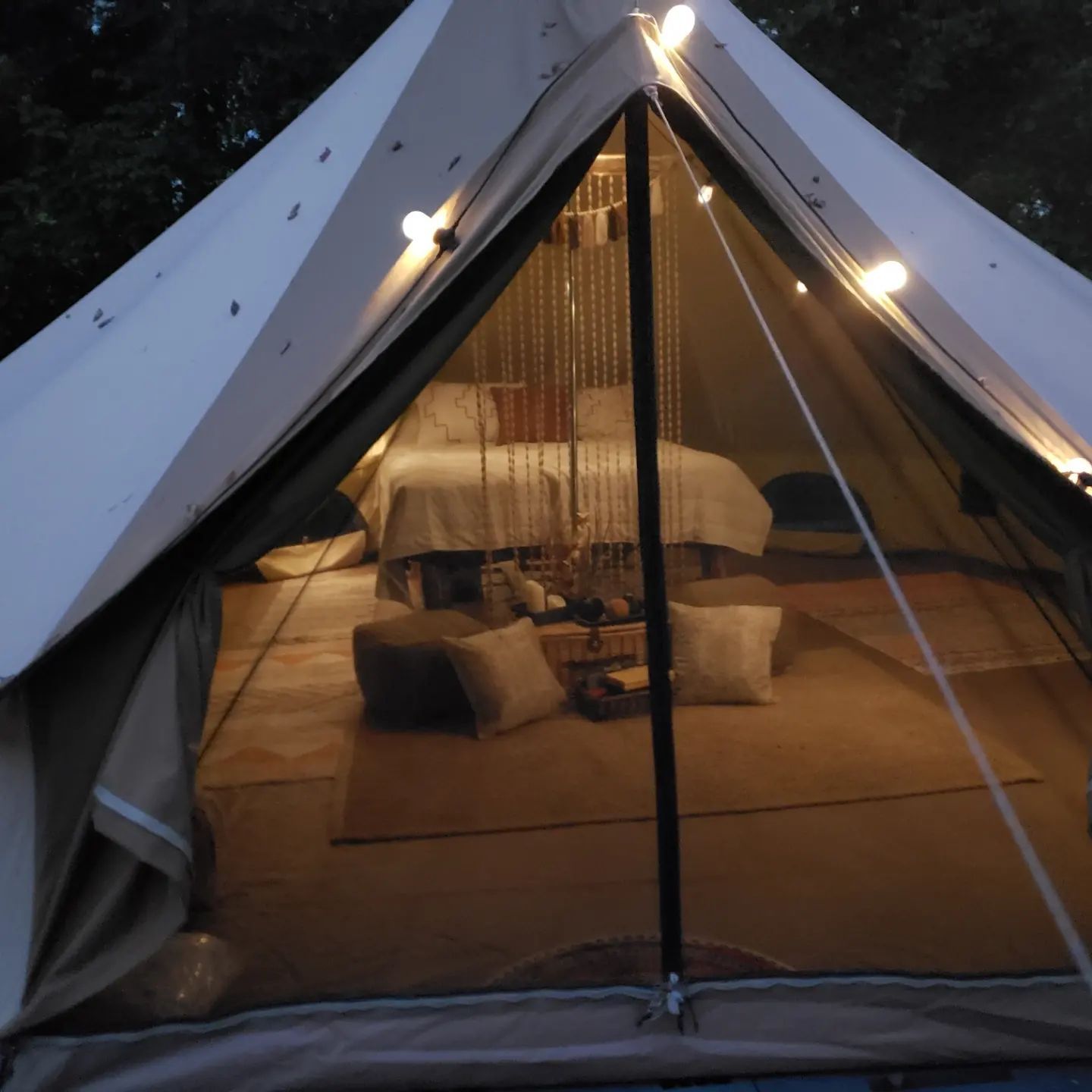Glowing tent interior at dusk with cozy bed, pillows, and string lights inside a white canvas tent