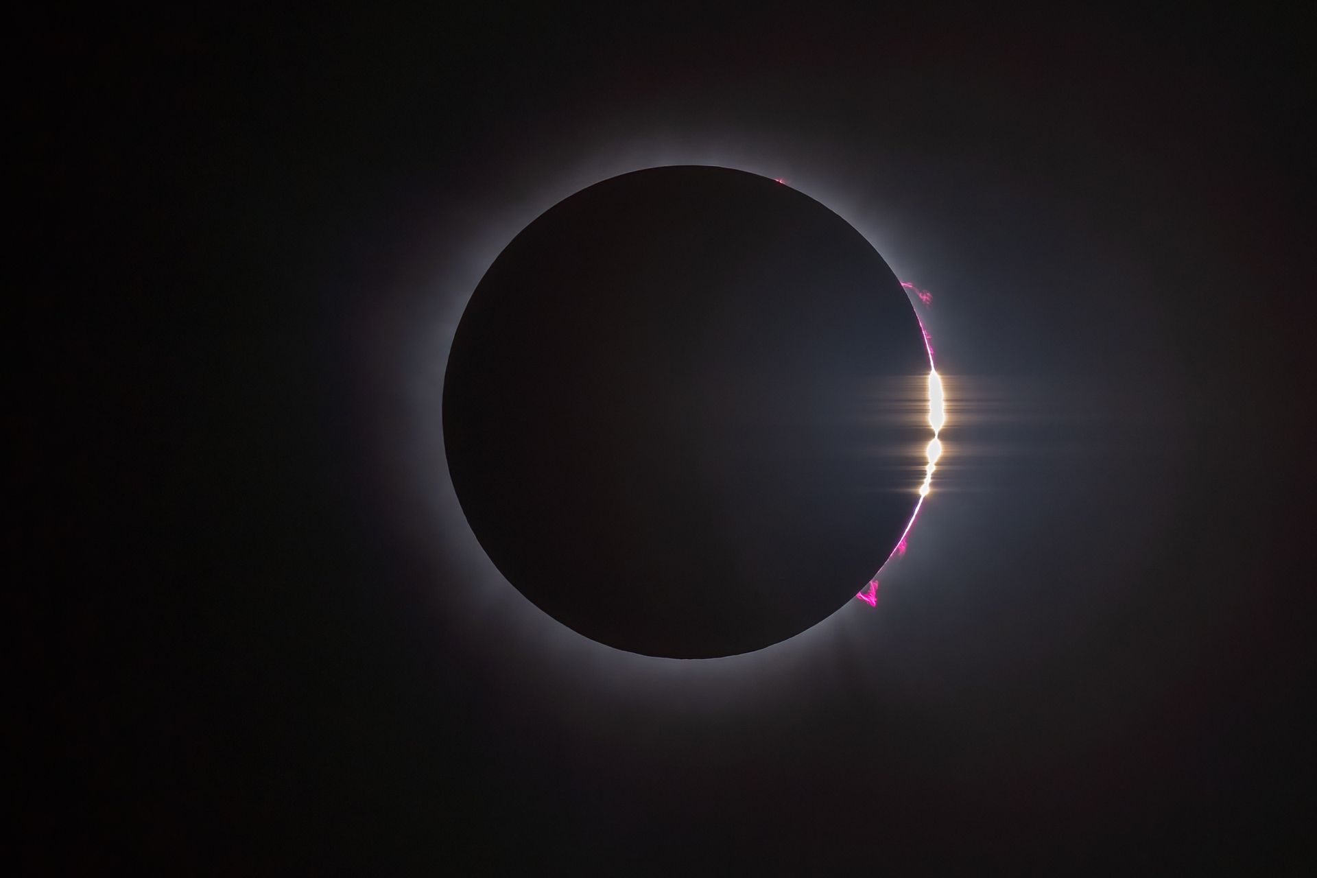 Total solar eclipse with the sun’s corona glowing behind a dark moon, and bright pink solar flares on the right.