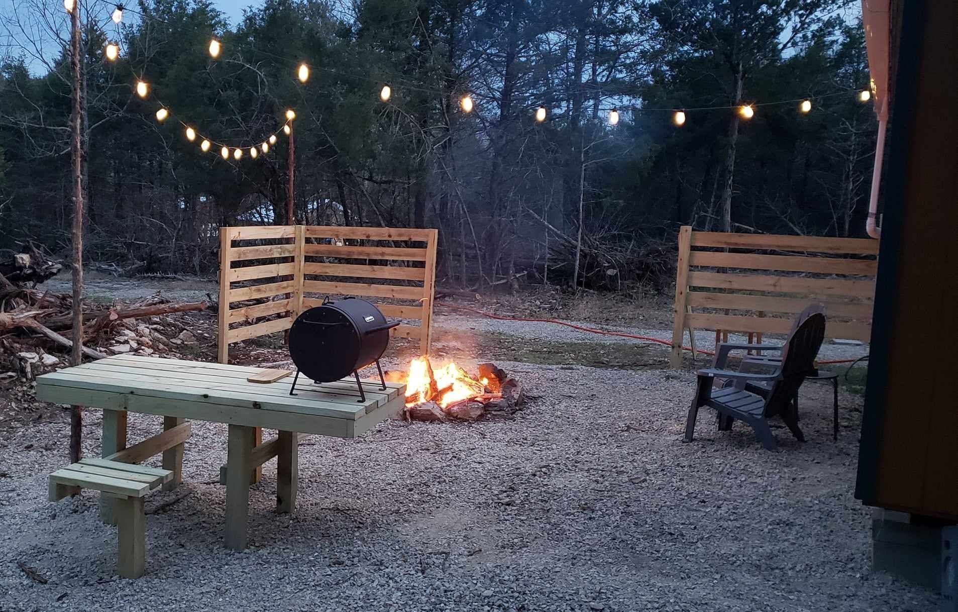 Backyard fire pit at dusk with string lights, benches, and gravel patio in a wooded setting