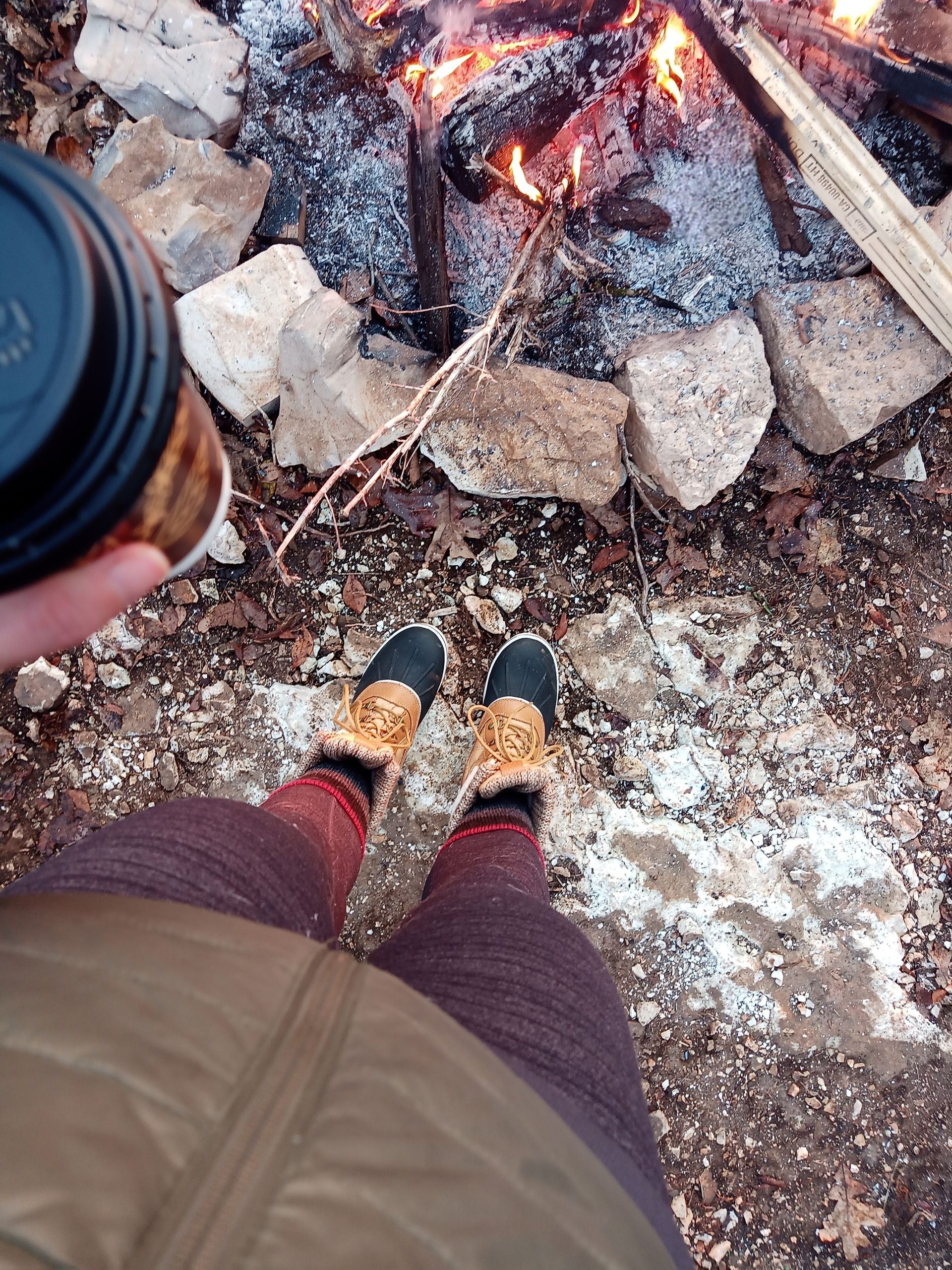 Person standing by a campfire, holding a coffee cup, wearing boots near stones and snow-covered ground.