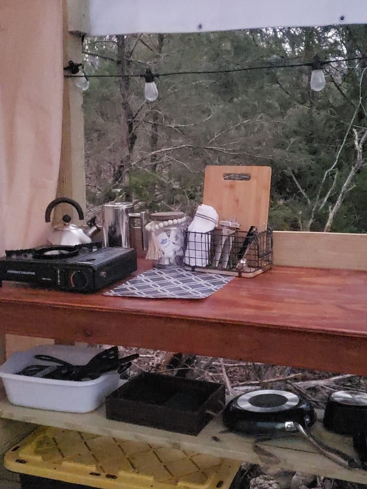 Outdoor wooden table with teapot, basket, and camping gear under a canopy in a wooded area