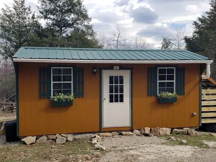 Small orange cabin with green metal roof and white door, set among trees and rocks.