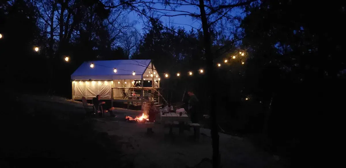 Lit tent at night with string lights and a small campfire in a wooded outdoor setting