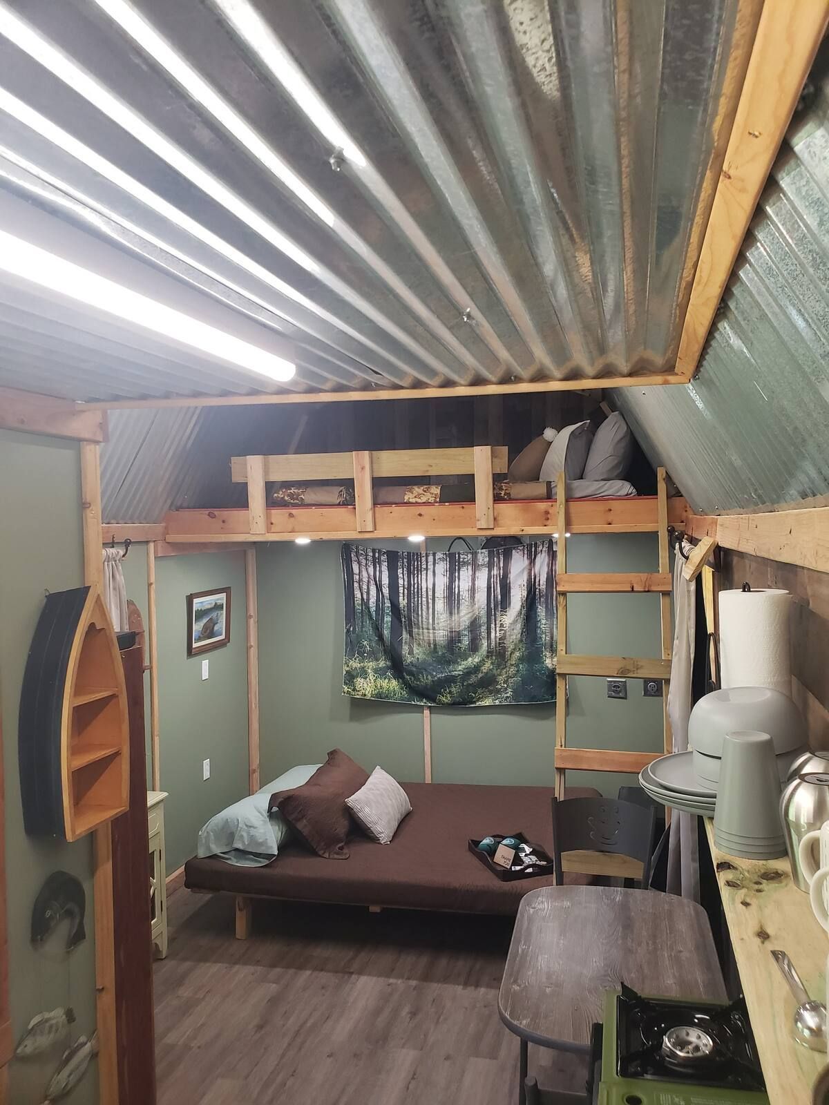 Tiny rustic loft room with a bed, ladder to a sleeping loft, corrugated metal ceiling, and forest tapestry.