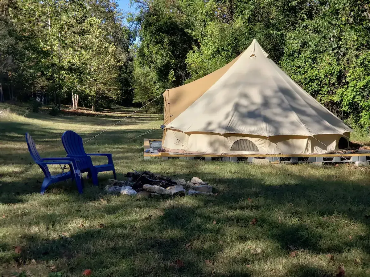 Beige canvas tent in a grassy campsite with blue chairs and a fire ring among trees.