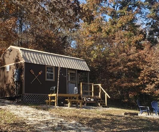Small cabin with a porch in a wooded autumn setting, with two blue chairs nearby.